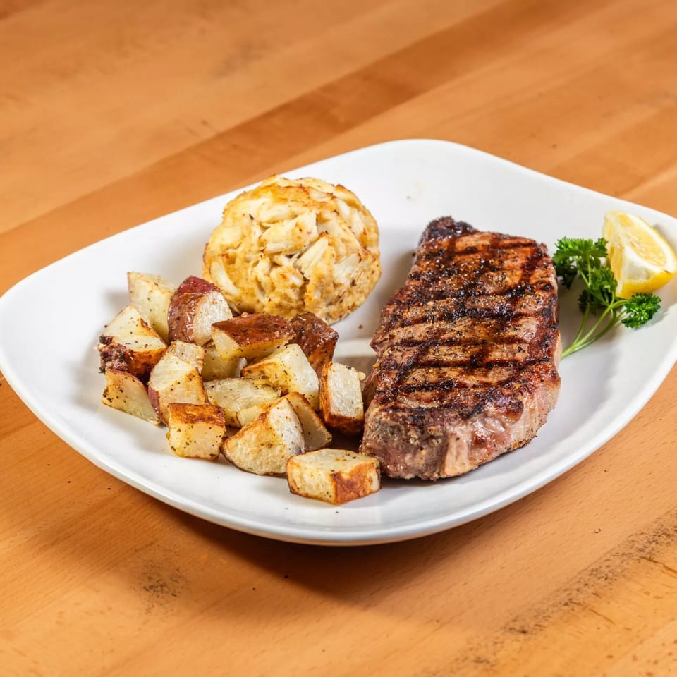 Grilled steak, crab cake, roasted potatoes, parsley, and lemon wedge on a white plate.