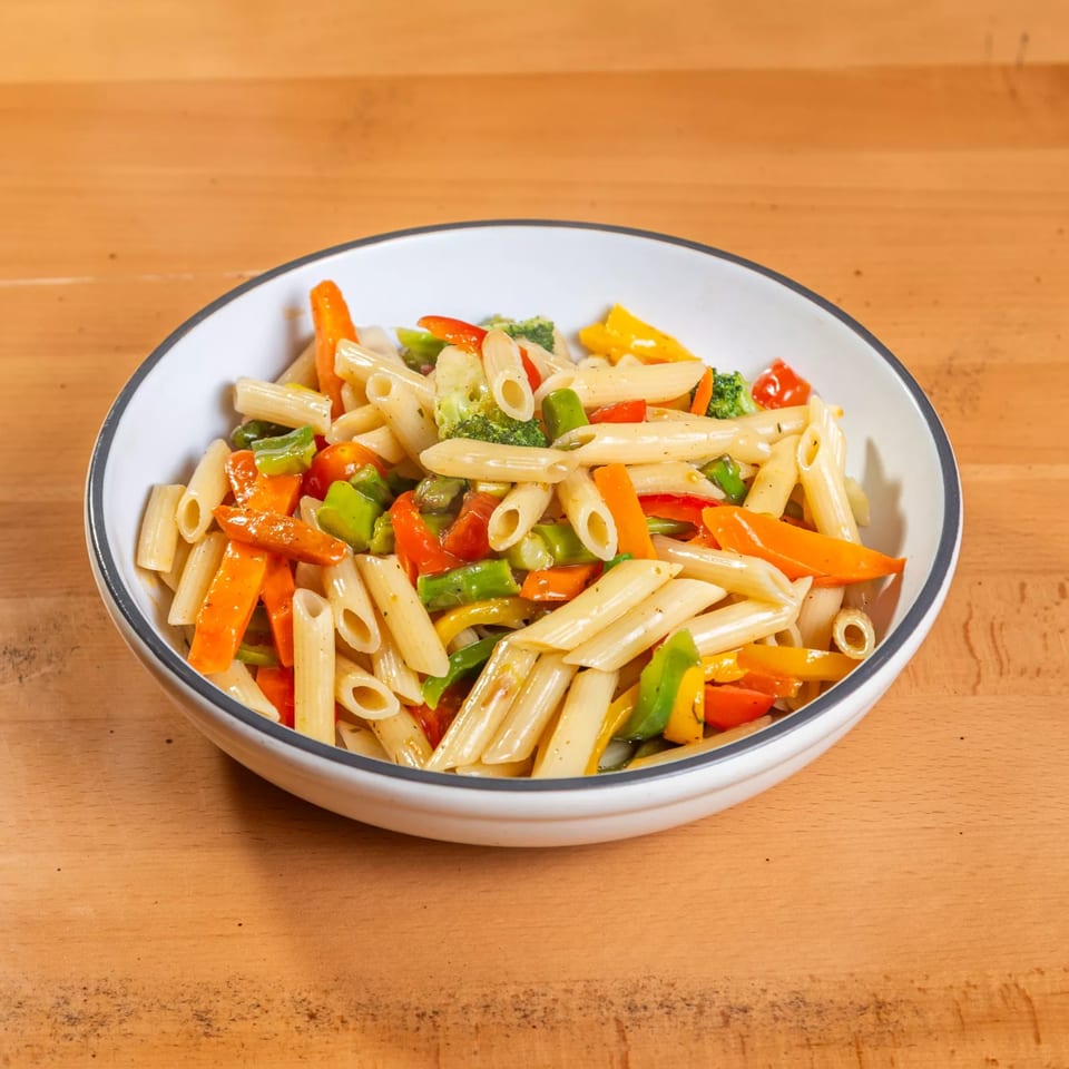 A white bowl filled with Rincon Italiano's penne pasta, colorful bell peppers, and broccoli on a wooden table.