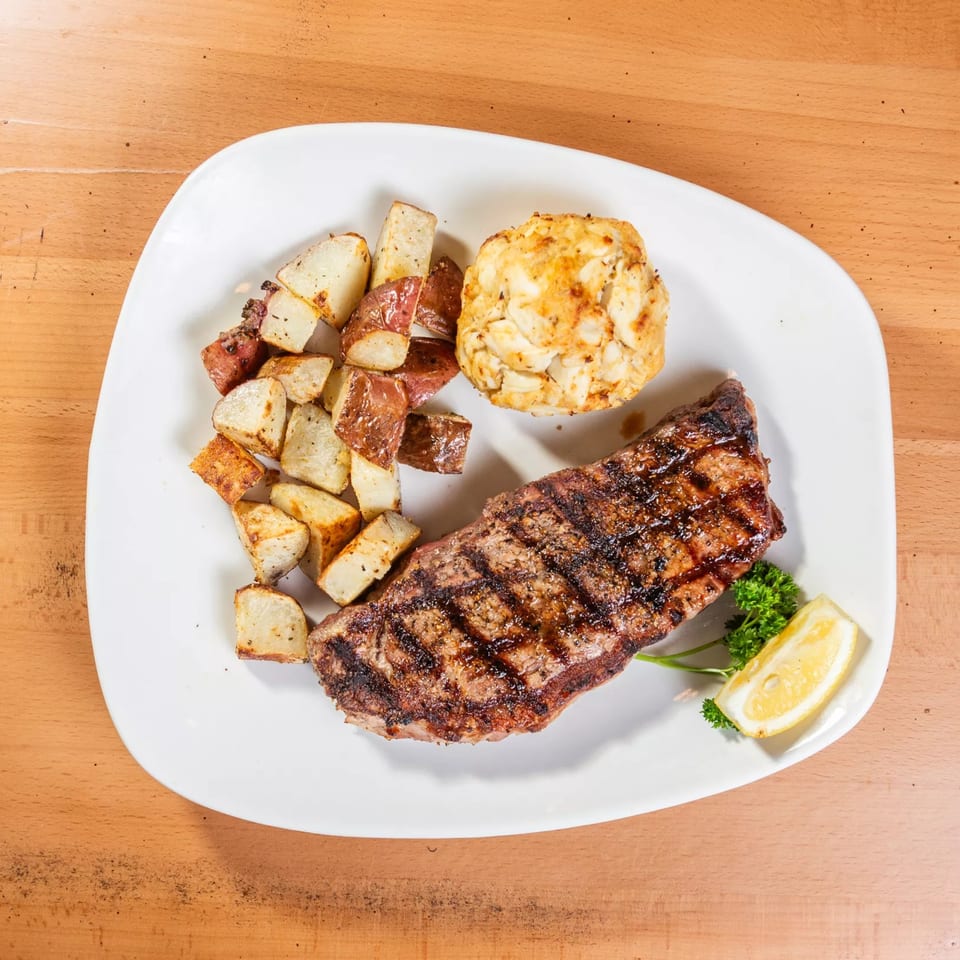 Grilled steak, roasted potatoes, and a crab cake with parsley and lemon on a white plate.